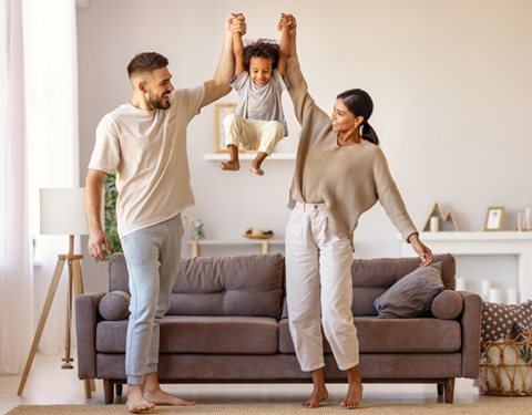 a man and woman standing in a living room with a child jumping on a couch